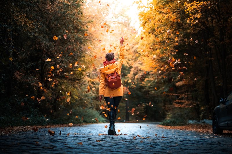 Donna con impermeabile giallo che cammina su una strada on foglie che le cadono attorno